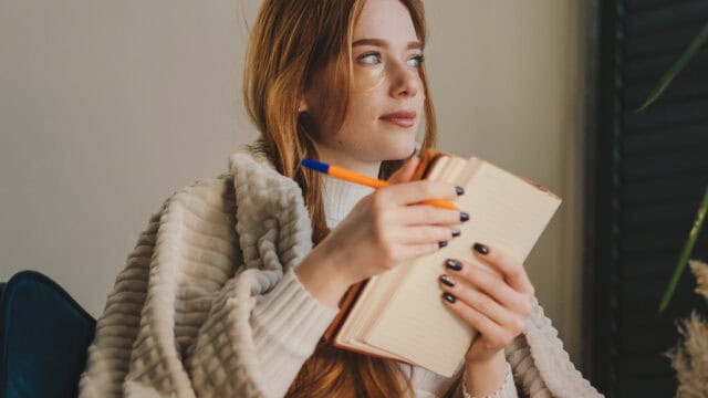 A woman with long red hair, wrapped in a beige blanket, holds a pen and notebook while looking thoughtfully to the side.