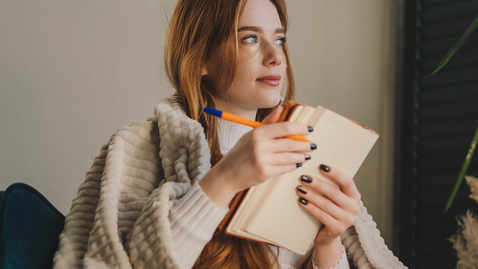 A woman with long red hair, wrapped in a beige blanket, holds a pen and notebook while looking thoughtfully to the side.