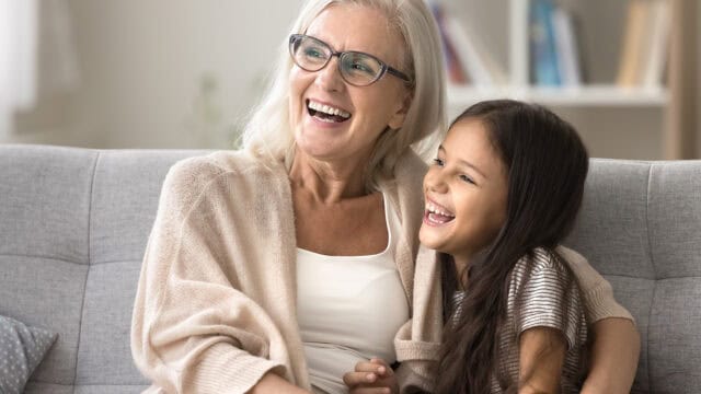 An older woman with gray hair and glasses sits on a couch, smiling and laughing with a young girl with long brown hair. They are hugging and appear happy in a cozy living room.