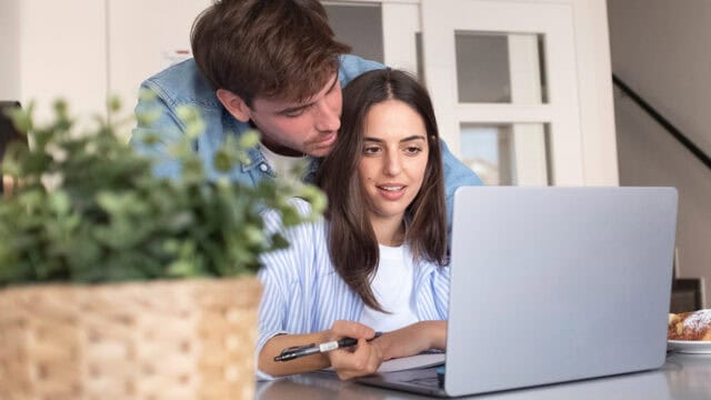 A man leans over a womans shoulder as they look at a laptop together. She holds a pen and has papers nearby. A potted plant is in the foreground.
