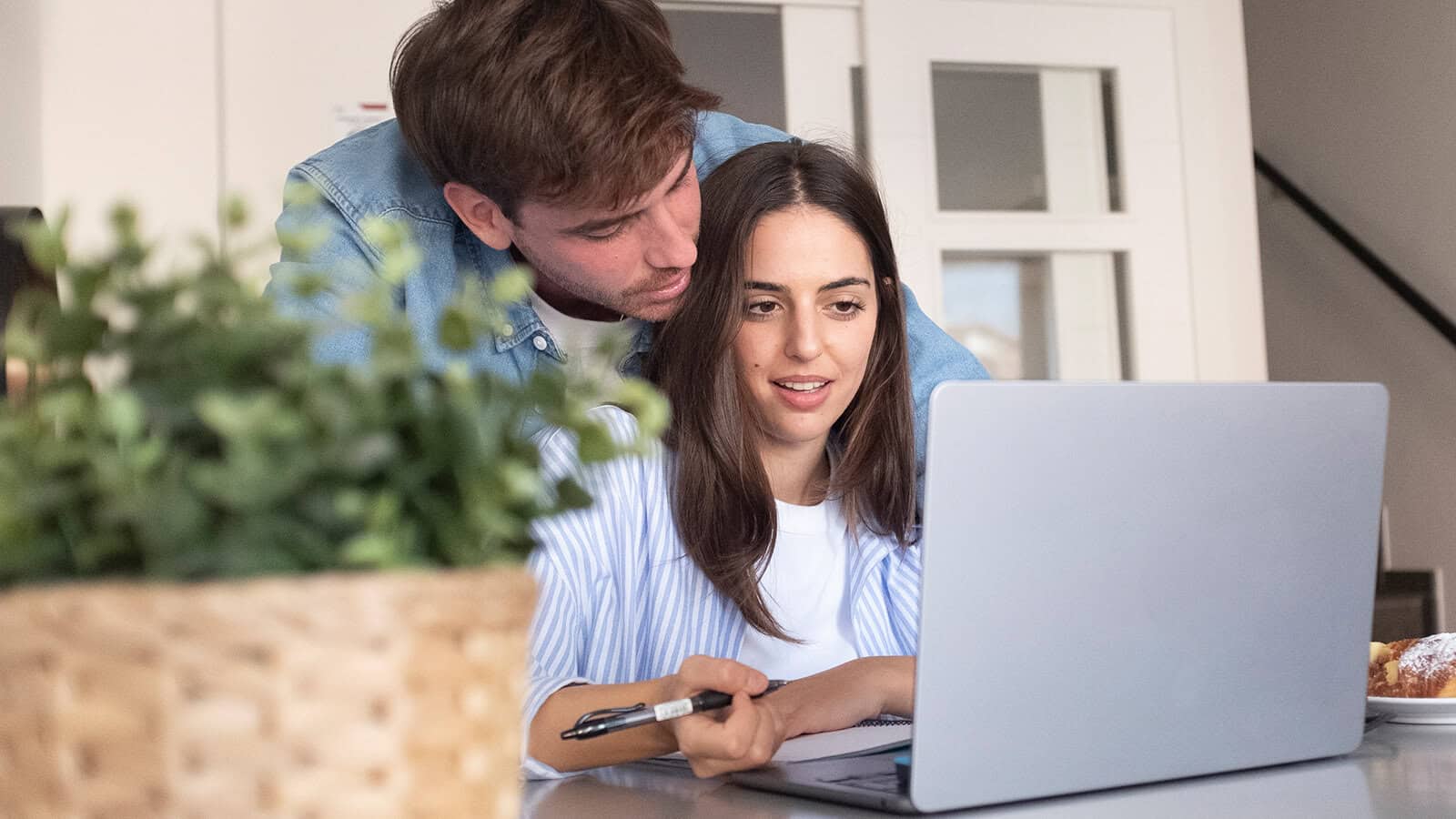 A man leans over a womans shoulder as they look at a laptop together. She holds a pen and has papers nearby. A potted plant is in the foreground.