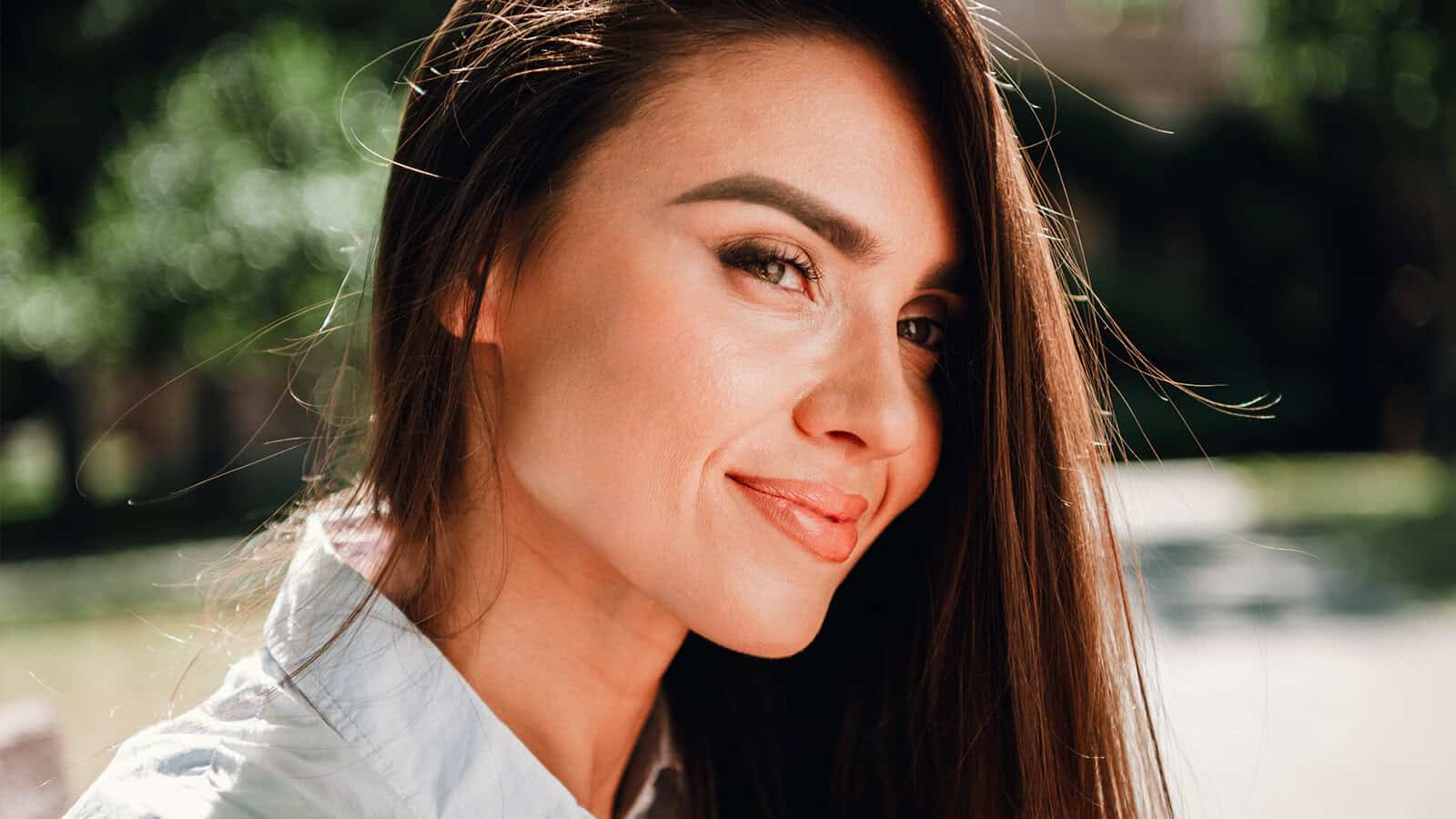 A woman with long brown hair and light makeup smiles while looking slightly over her shoulder outdoors on a sunny day.