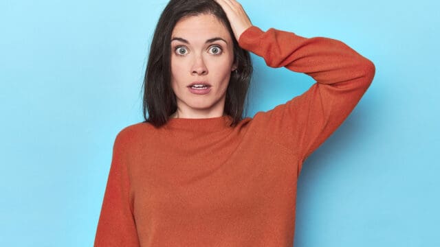 A person with long dark hair, wearing an orange sweater, looks surprised with wide eyes and mouth slightly open, holding one hand to the side of their head, standing against a light blue background.