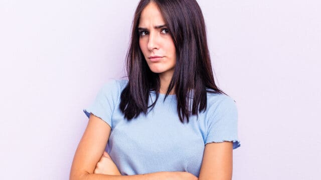Woman with long dark hair and a blue short-sleeve shirt stands with arms crossed, looking forward with a skeptical or annoyed expression. Pale purple background.