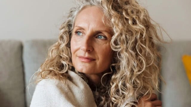 A woman with curly blonde hair and blue eyes looks thoughtfully to the side while sitting indoors, wrapped in a light-colored sweater.