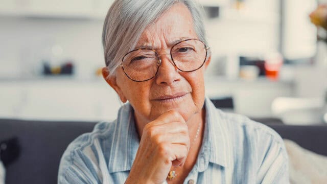 Older woman with gray hair and glasses sits indoors, wearing a striped shirt. She looks thoughtfully at the camera, resting her chin on her hand. The background is softly blurred.