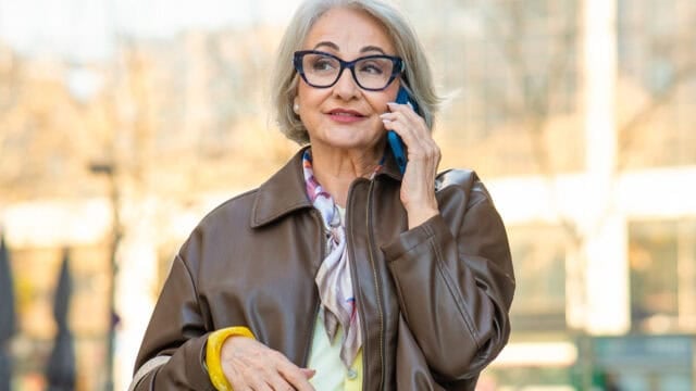 Older woman with gray hair and glasses, wearing a brown jacket and scarf, talks on a smartphone outdoors.