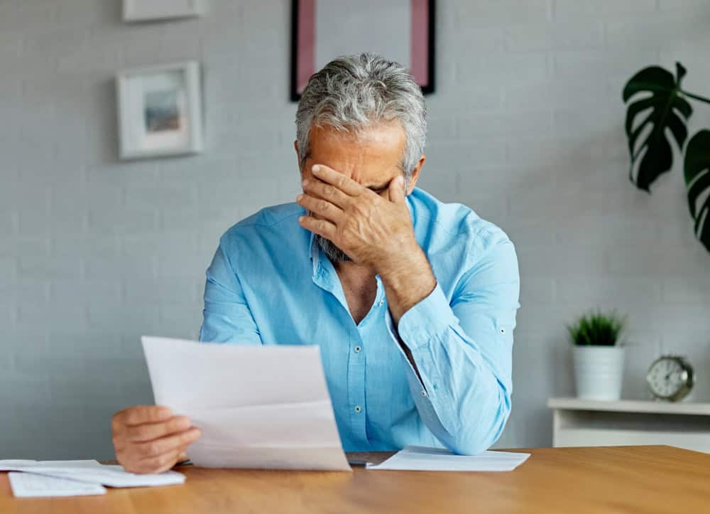 A millennial man in a blue shirt sits at a table, holding papers in one hand and covering his face with the other, appearing stressed or worried. There are plants, a clock, and framed photos in the background.