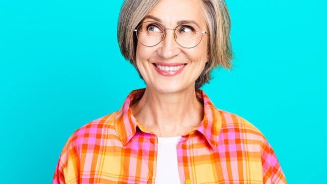 Smiling older woman with short gray hair and glasses, wearing an orange and pink plaid shirt, looking to the side against a bright blue background.
