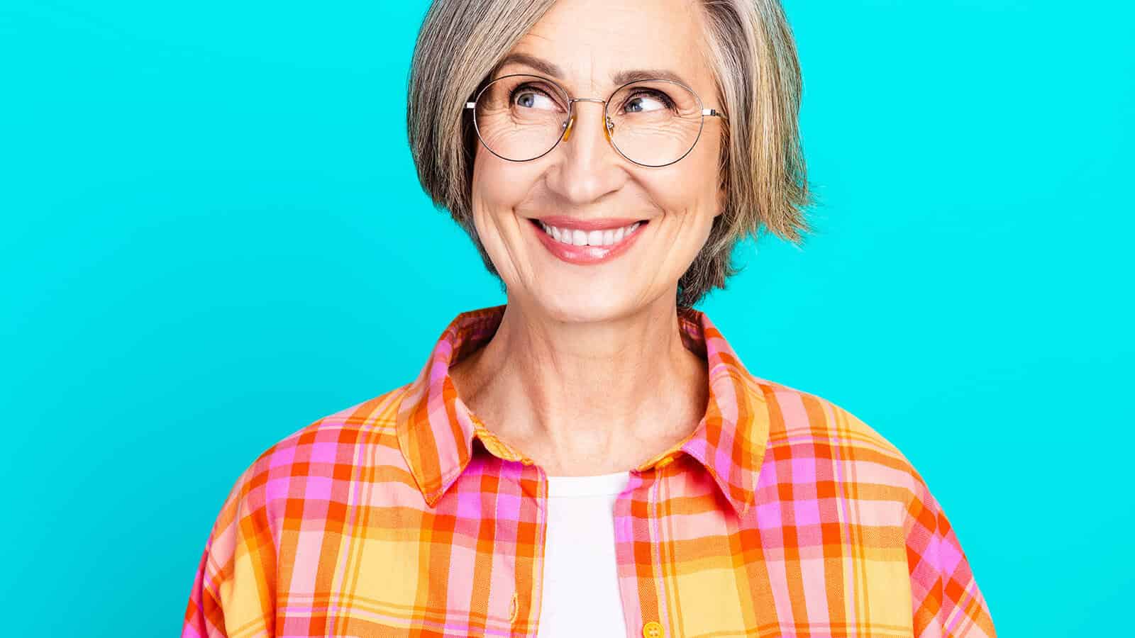 Smiling older woman with short gray hair and glasses, wearing an orange and pink plaid shirt, looking to the side against a bright blue background.