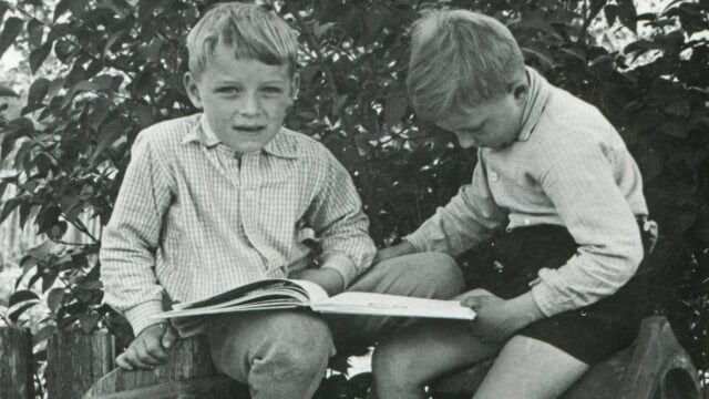 Two young boys sit outside on a wooden fence, one reading a book while the other looks down at the pages. Trees with leafy branches are in the background. Both boys wear long-sleeve shirts and shorts.