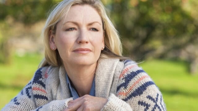 A woman with blonde hair wearing a patterned sweater sits outdoors on a sunny day, looking thoughtfully into the distance with a slight smile. The background is blurred with green trees and grass.