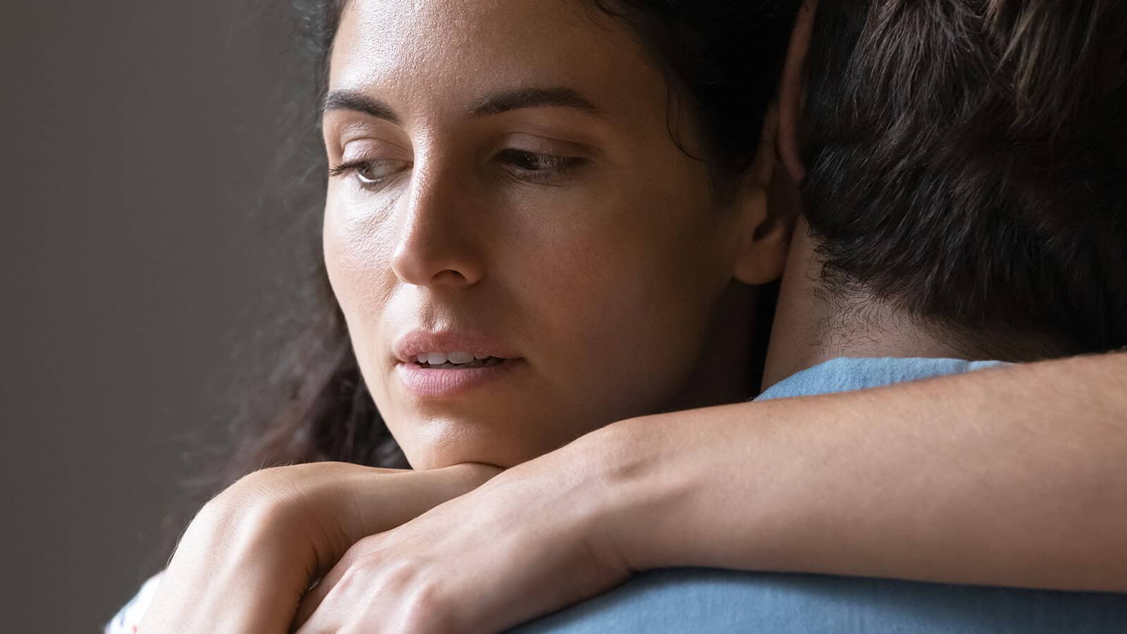 A woman with a concerned expression embraces a man, her arms resting on his shoulders while she looks away, appearing thoughtful or worried.