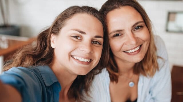 Two women smiling and taking a close-up selfie together indoors, both wearing blue shirts and appearing happy.
