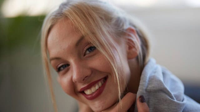 A young woman with blonde hair and blue eyes smiles warmly at the camera, resting her chin on her hands. She wears a light gray hoodie and has dark red lipstick and nail polish. The background is softly blurred.