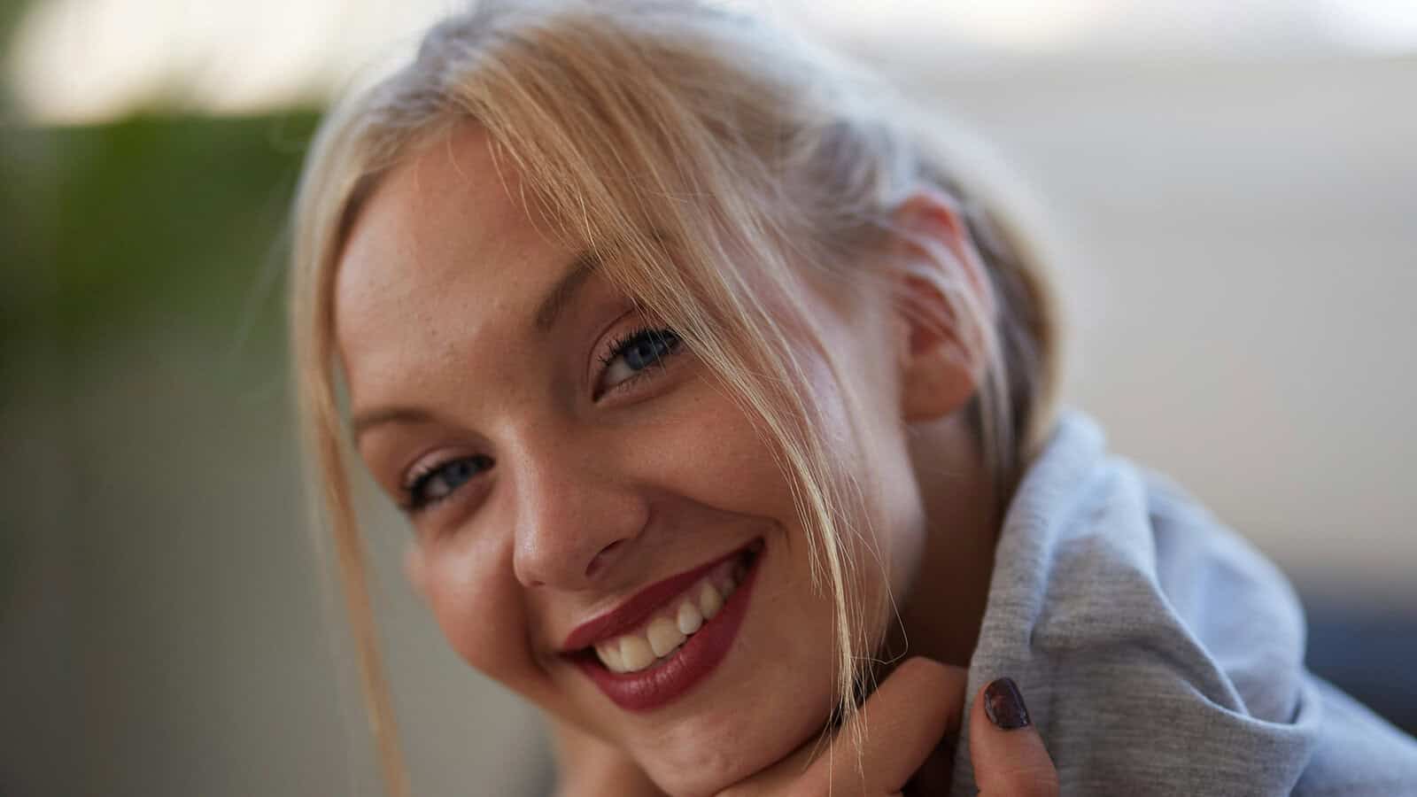 A young woman with blonde hair and blue eyes smiles warmly at the camera, resting her chin on her hands. She wears a light gray hoodie and has dark red lipstick and nail polish. The background is softly blurred.
