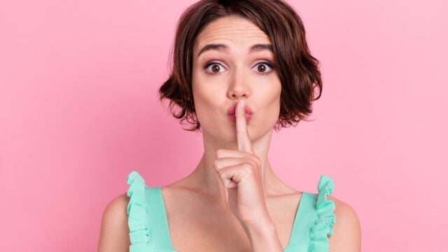 A woman with short brown hair in a mint green top holds her finger to her lips, signaling shh against a pink background.
