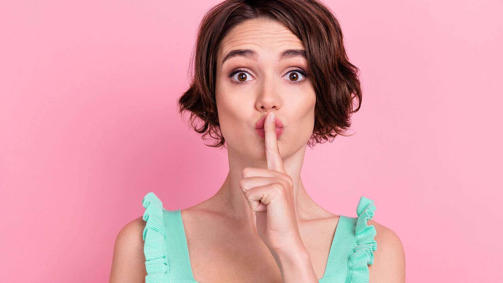 A woman with short brown hair in a mint green top holds her finger to her lips, signaling shh against a pink background.
