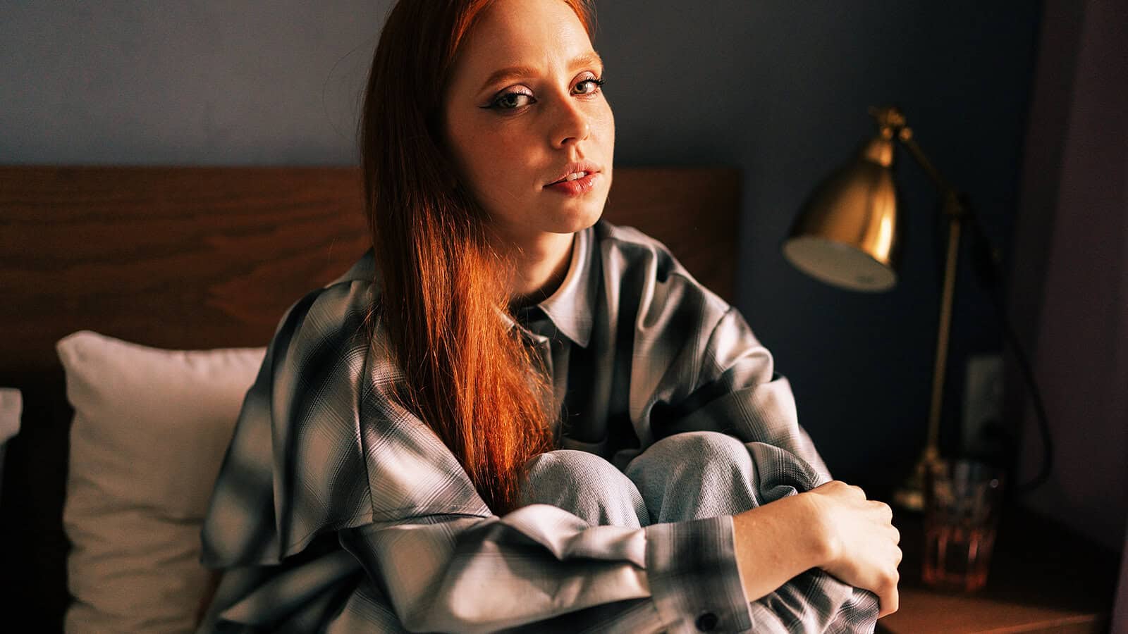 A woman with long red hair sits on a bed, hugging her knees and looking at the camera. She wears a plaid shirt and is beside a gold lamp on a wooden nightstand. The lighting is warm and soft.