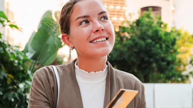 A woman in a tan blazer and white shirt smiles while holding a credit card outdoors, with green plants and blurred buildings in the background.