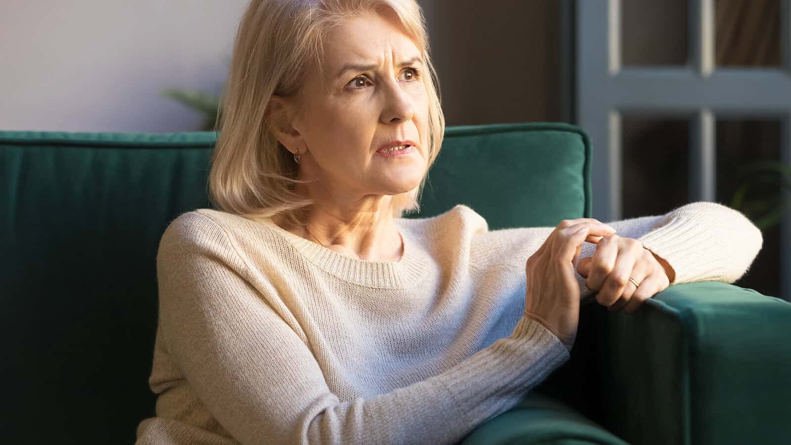 Older woman with short blonde hair sits on a green sofa, looking concerned or thoughtful, with her hands resting on the arm of the couch in soft indoor lighting.