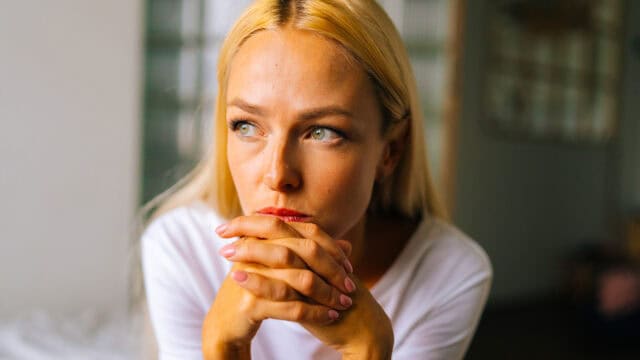 A woman with long blonde hair rests her chin on her clasped hands, looking thoughtfully to the side. She is indoors, wearing a white shirt, with a soft, unfocused background.