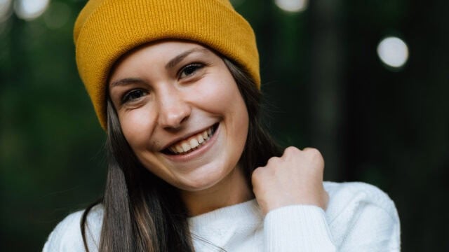 A young woman smiles warmly at the camera, wearing a yellow knit beanie and a white sweater, with a blurred green outdoor background.