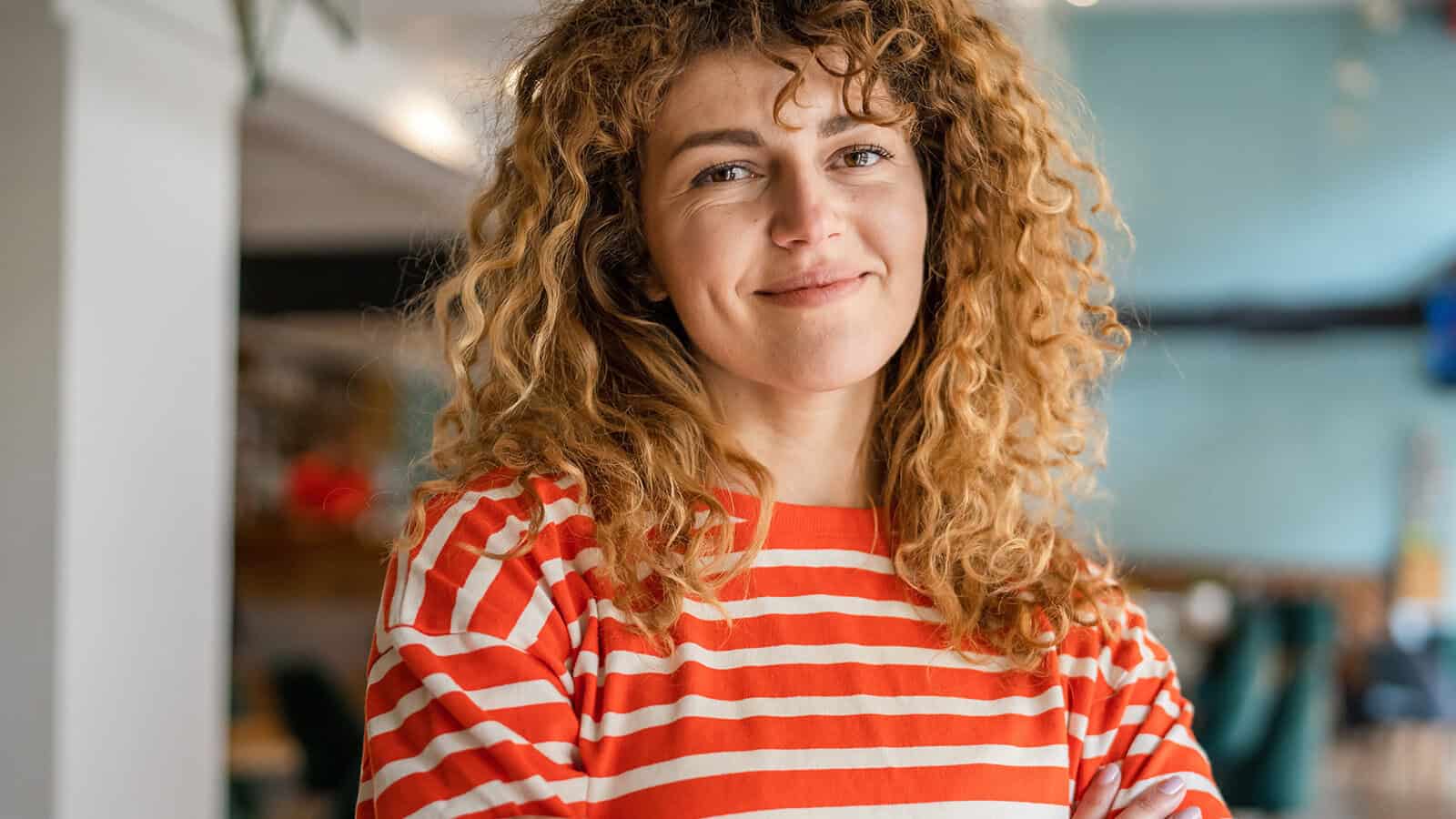 A person with curly hair wearing a red and white striped shirt smiles softly, standing indoors with a blurred background.