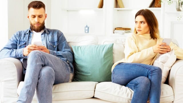 A man and woman sit on a couch, both looking at their phones. The woman looks at the man with a serious expression, while the man looks down at his phone. Both appear distant and unhappy.