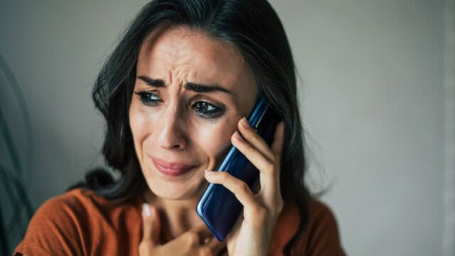 A woman with long dark hair holds a phone to her ear, looking distressed and worried. She touches her chest with her hand, and tears are visible on her face.