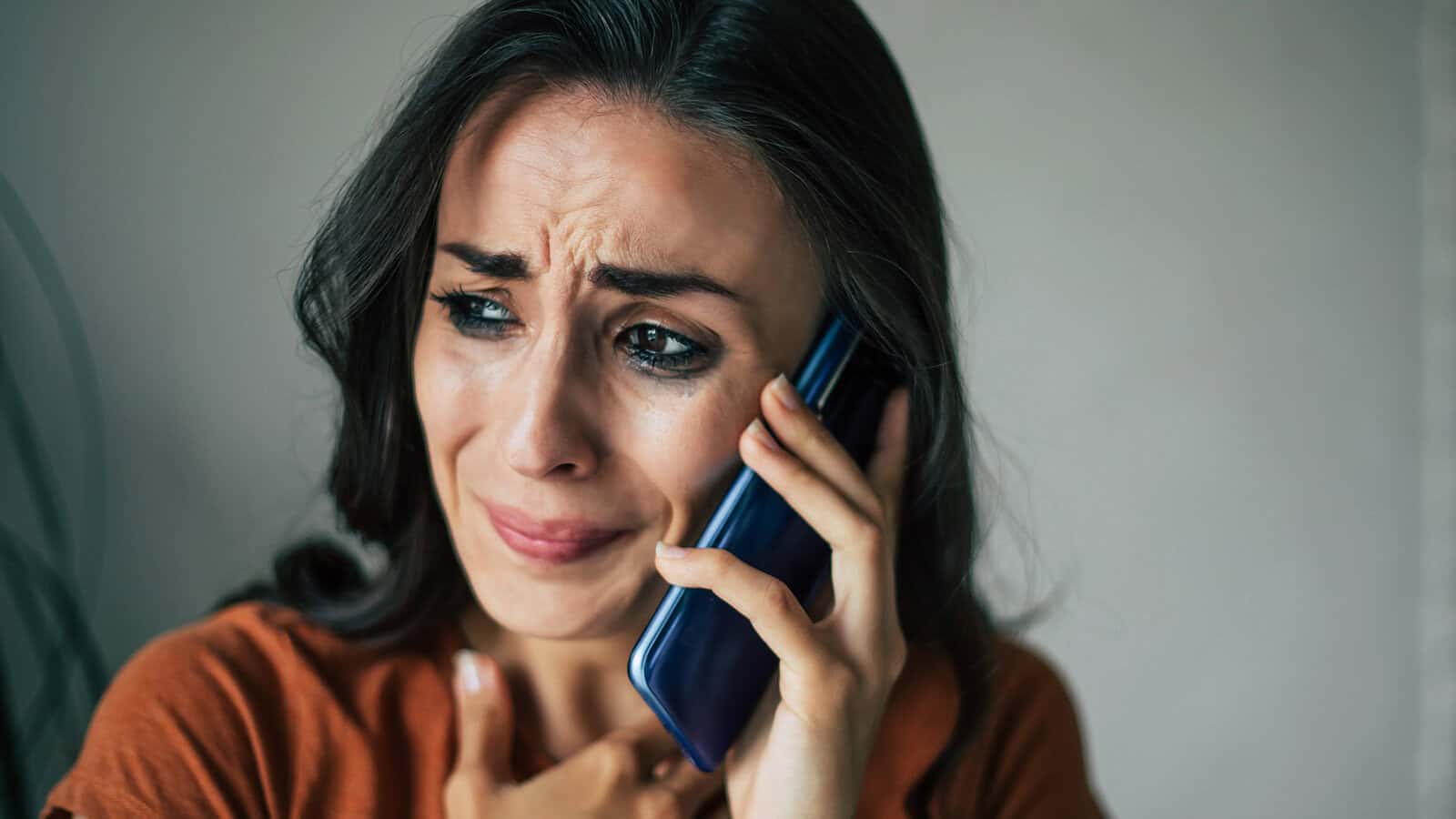 A woman with long dark hair holds a phone to her ear, looking distressed and worried. She touches her chest with her hand, and tears are visible on her face.