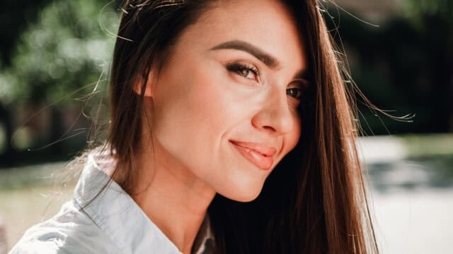A woman with long brown hair and a white shirt smiles softly outdoors, with sunlight highlighting her face and a blurred green background.