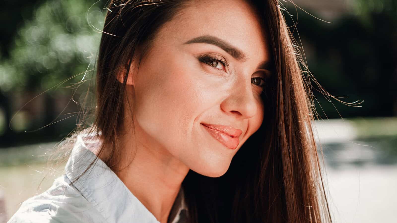 A woman with long brown hair and a white shirt smiles softly outdoors, with sunlight highlighting her face and a blurred green background.