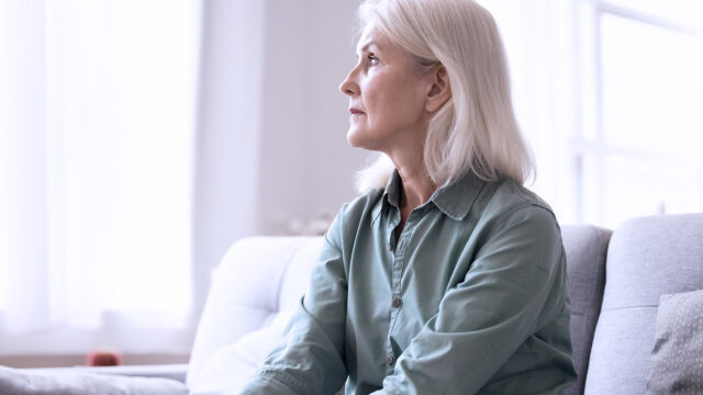 An older woman with gray hair sits on a light-colored sofa, looking thoughtfully out a bright window. She wears a green button-up shirt and appears calm and reflective.