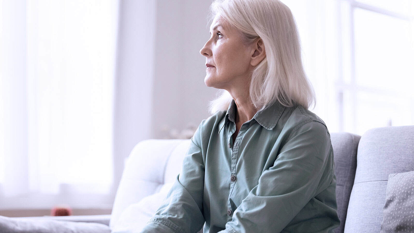 An older woman with gray hair sits on a light-colored sofa, looking thoughtfully out a bright window. She wears a green button-up shirt and appears calm and reflective.