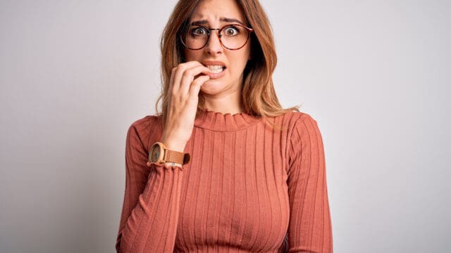 A woman with long hair and glasses wearing a ribbed, long-sleeve shirt looks anxious, biting her nails, and staring forward against a plain, light background.
