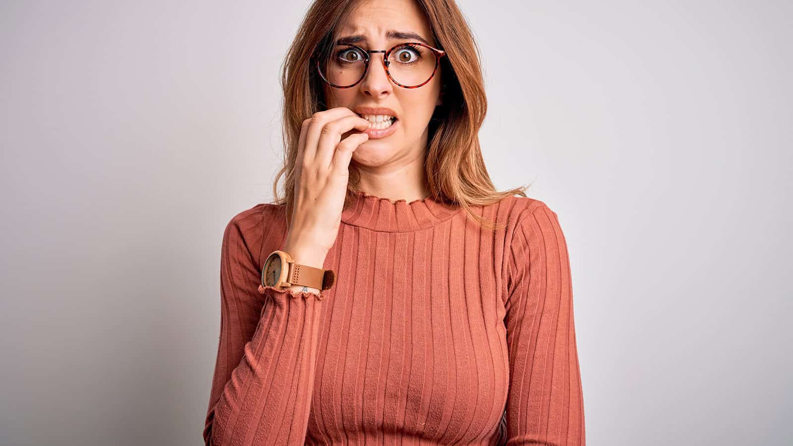 A woman with long hair and glasses wearing a ribbed, long-sleeve shirt looks anxious, biting her nails, and staring forward against a plain, light background.