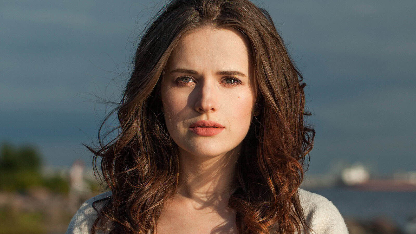 A woman with long wavy brown hair stands outdoors, looking directly at the camera, with a soft natural background and sunlight illuminating her face, subtly reflecting on the small behaviors that shape relationships.