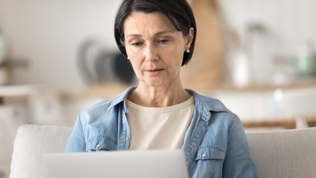 Middle-aged woman with short dark hair wearing a denim shirt and beige top sits on a light sofa, looking thoughtfully at a laptop screen in a bright, softly focused room.
