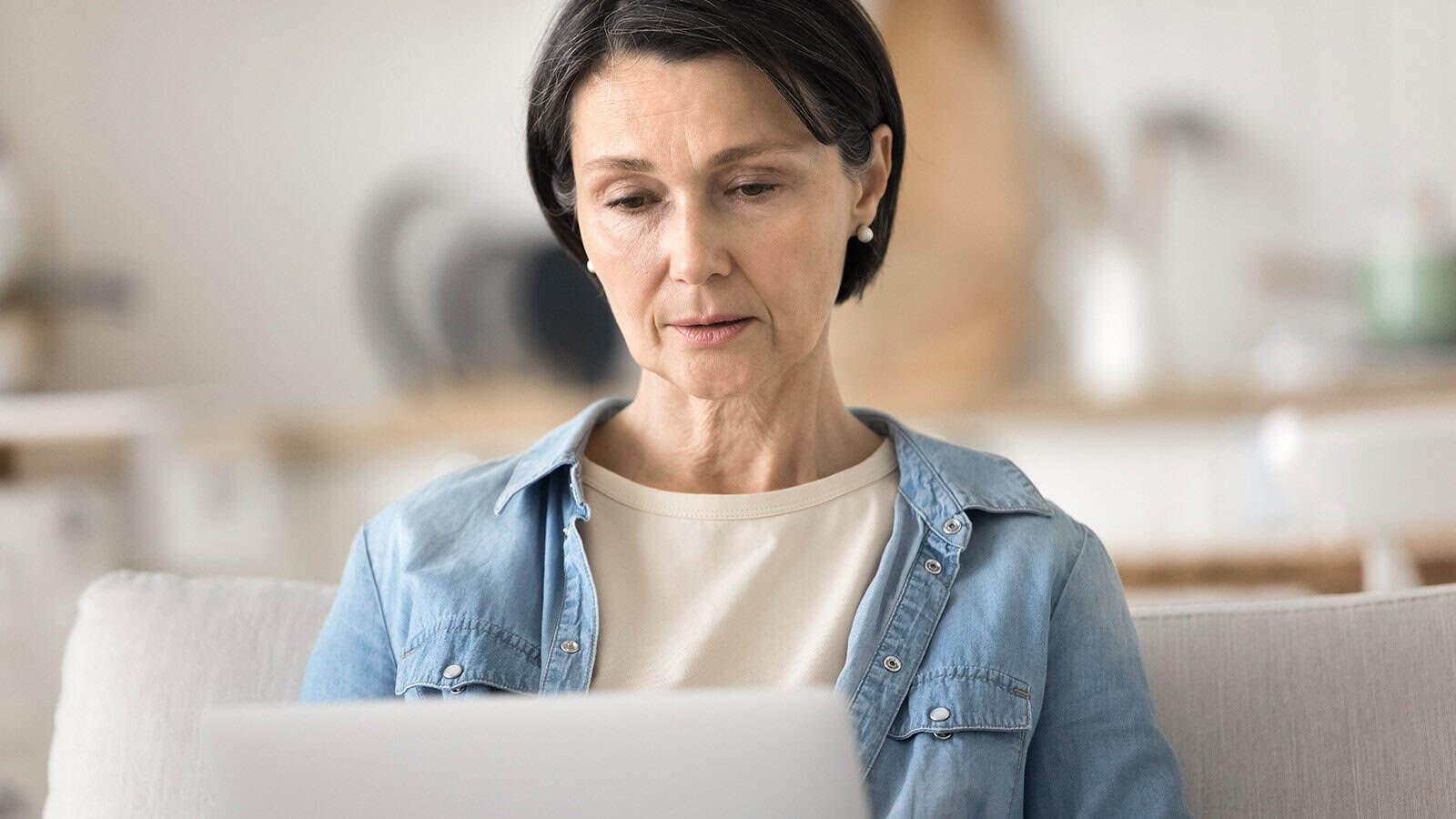 Middle-aged woman with short dark hair wearing a denim shirt and beige top sits on a light sofa, looking thoughtfully at a laptop screen in a bright, softly focused room.