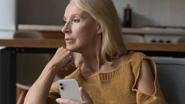 A woman with blonde hair wearing a mustard sweater sits indoors, holding a smartphone and looking thoughtfully out of the frame.