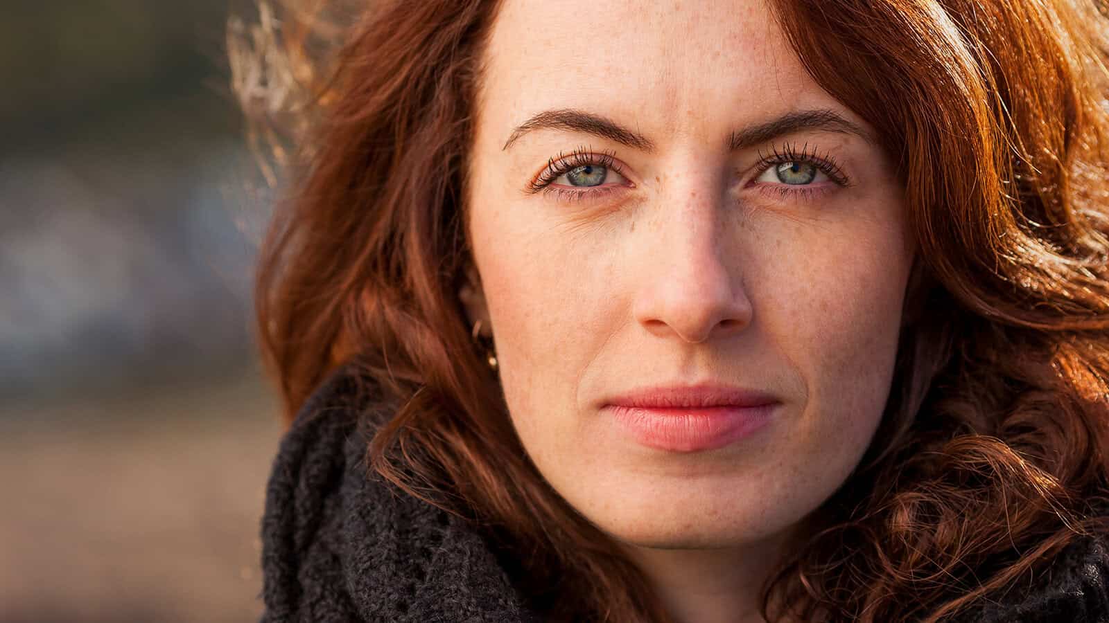 A woman with long red hair and green eyes looks directly at the camera, wearing a dark scarf and an outdoor background blurred behind her.