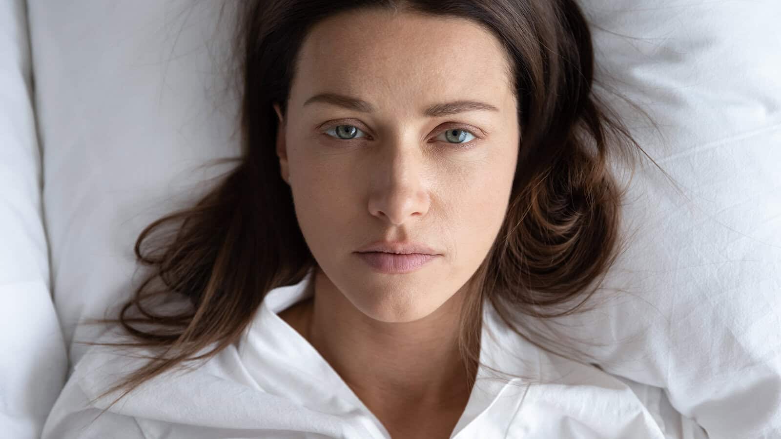 A woman with straight brown hair lies on her back in bed, head on a white pillow, looking directly at the camera with a calm, neutral expression. She is wearing a white top.
