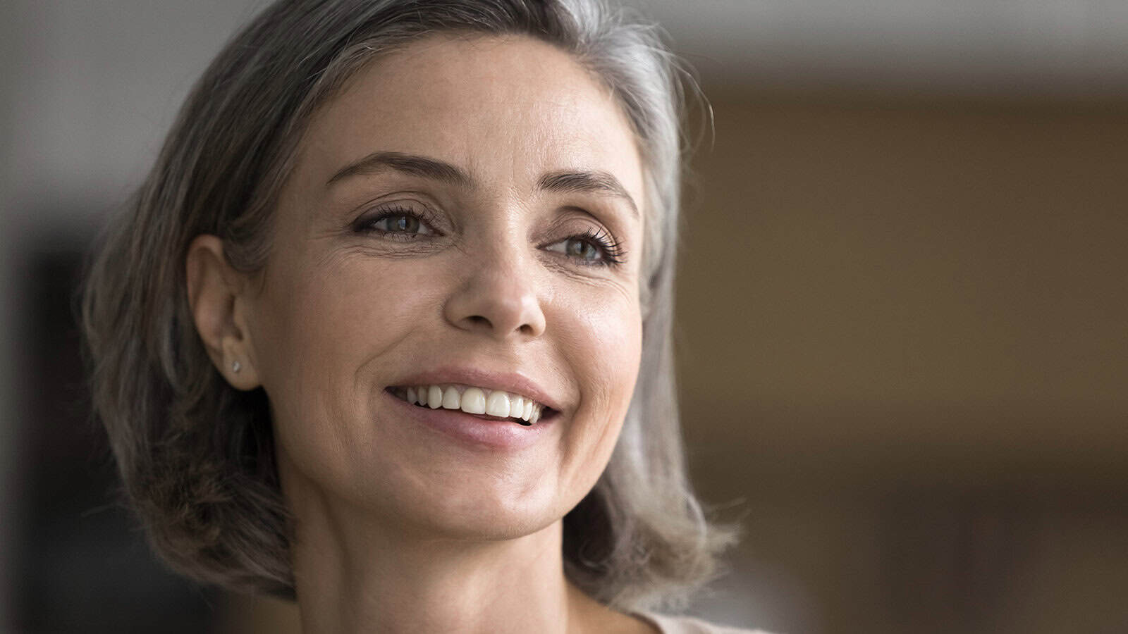 A woman with short gray hair smiles gently, looking slightly to the side. She has light skin, subtle makeup, and wears small stud earrings. The background is softly blurred, highlighting her face.