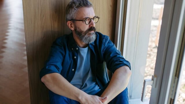 A man with glasses and a gray beard sits on the floor by a window, looking outside thoughtfully. He wears a blue shirt over a light t-shirt and jeans, with natural light coming through the window.