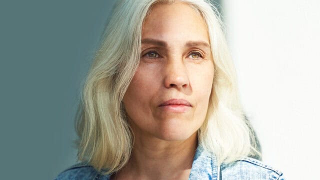 A woman with long gray hair and light skin gazes thoughtfully into the distance, illuminated by natural light, wearing a denim jacket—her expression quietly hinting at the feeling of aging and occasionally feeling out of place.