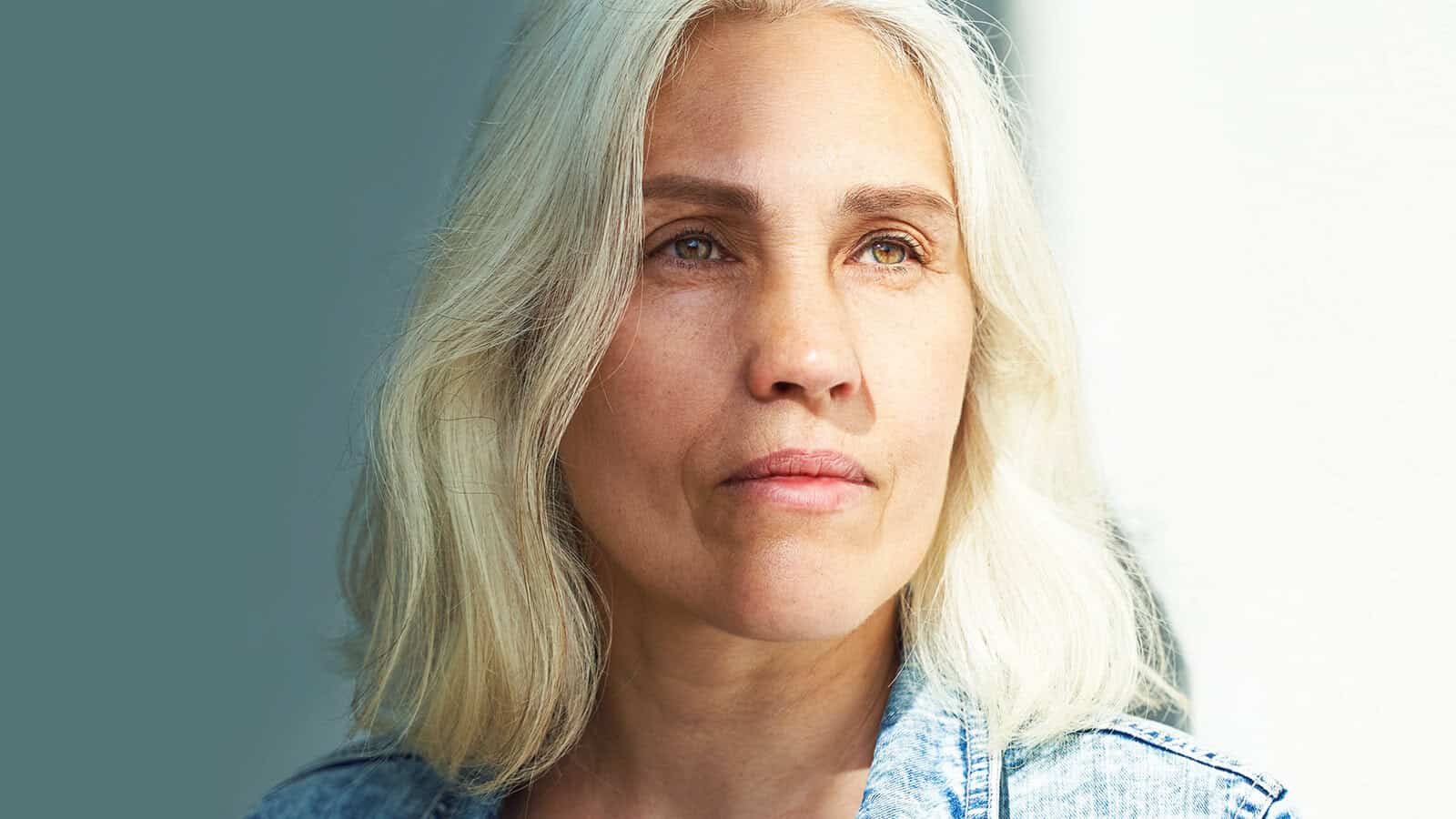 A woman with long gray hair and light skin gazes thoughtfully into the distance, illuminated by natural light, wearing a denim jacket—her expression quietly hinting at the feeling of aging and occasionally feeling out of place.