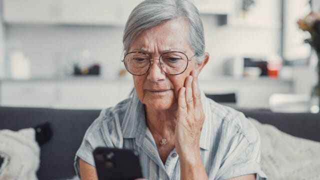 An older woman with gray hair and glasses looks concerned while holding her smartphone, touching her cheek as she sits on a couch in a well-lit living room.