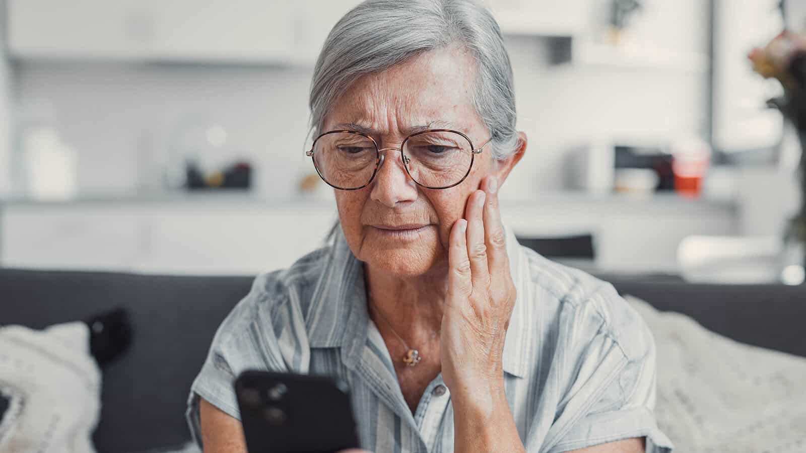 An older woman with gray hair and glasses looks concerned while holding her smartphone, touching her cheek as she sits on a couch in a well-lit living room.