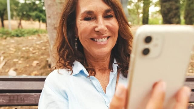 A smiling woman with brown hair sits on a bench outdoors, holding a smartphone and looking at its screen. Trees and greenery are visible in the blurred background.